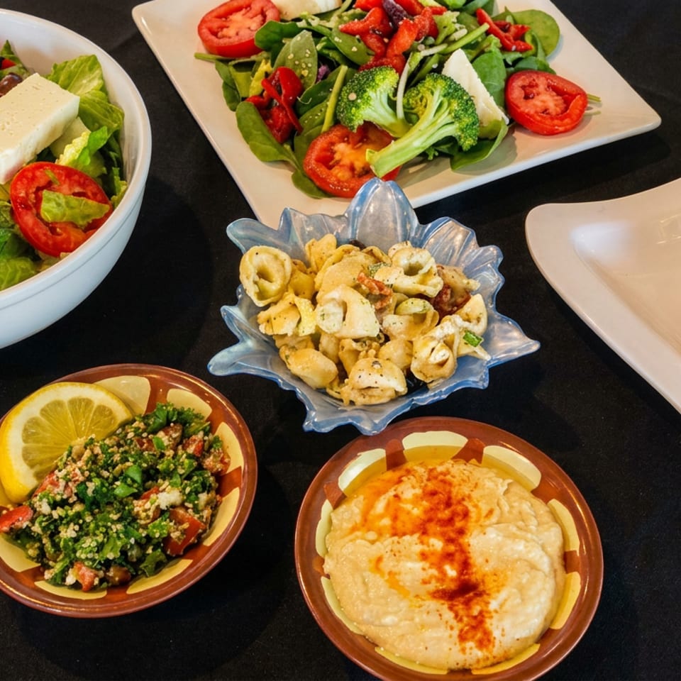 Assorted Mediterranean dishes, including salads, hummus, tabbouleh, pasta, and a pita filled with meat and vegetables, arranged on a table.