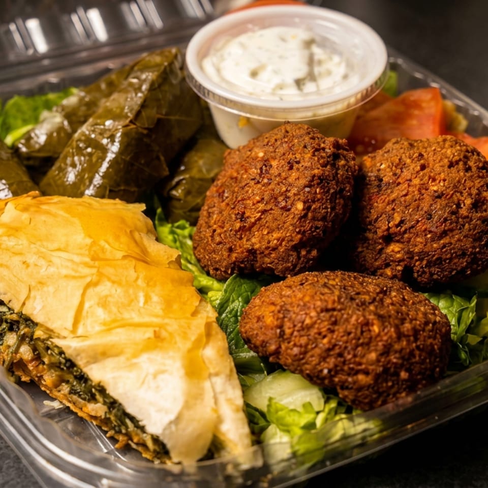 Mediterranean platter with falafel, stuffed grape leaves, spinach pastry, lettuce, tomatoes, and creamy dipping sauce in a takeaway container.