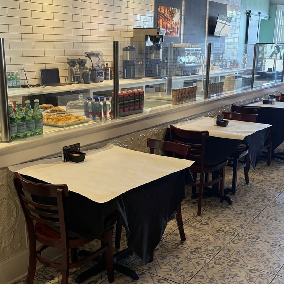 Bright restaurant dining room with white-tableclothed tables, patterned tile floors, counter service, drinks, and food displays.