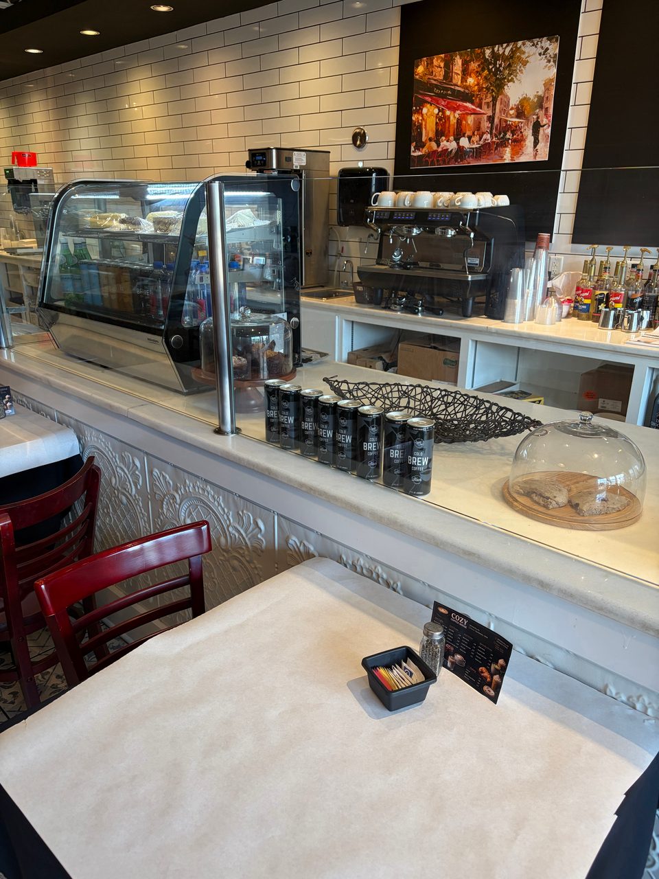 A cafe interior with a pastry display, coffee machine, cold brew cans, and a table with a menu.