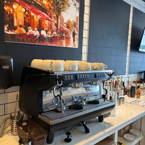 A large espresso machine with white cups on top, next to a painting of a cafe scene.