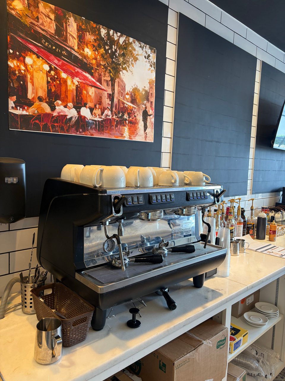 A black espresso machine with white cups on top, next to a painting of a Parisian cafe.