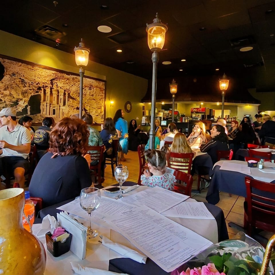 Busy restaurant dining room filled with patrons, tables set with menus and wine glasses, warm lighting, and decorative wall art.