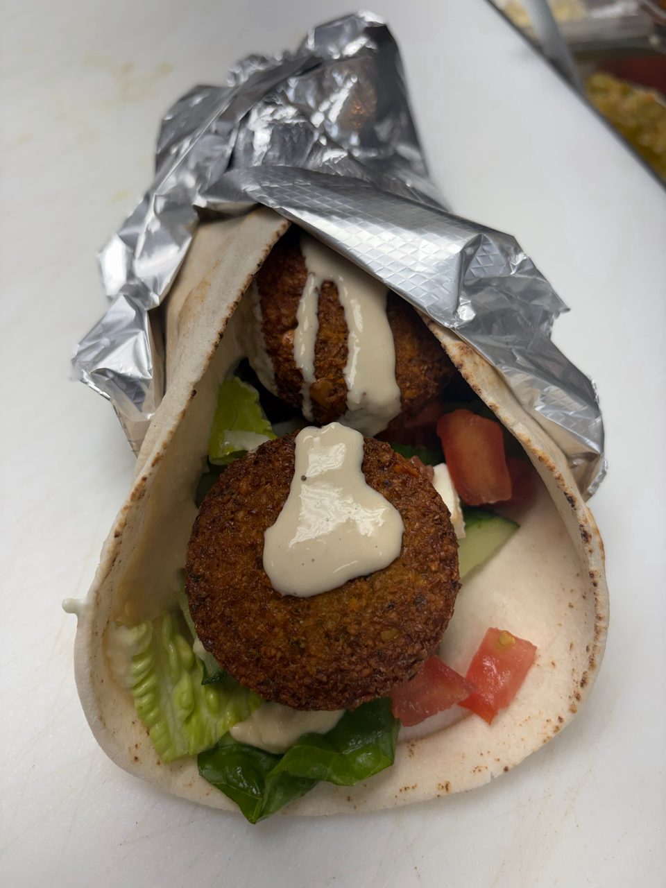 Falafel wrap with lettuce, tomatoes, cucumbers, and tahini sauce, partially wrapped in foil.