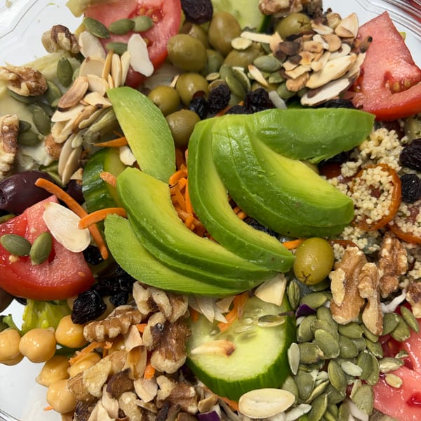 A vibrant salad in a clear container, featuring avocado, tomatoes, olives, nuts, and seeds.