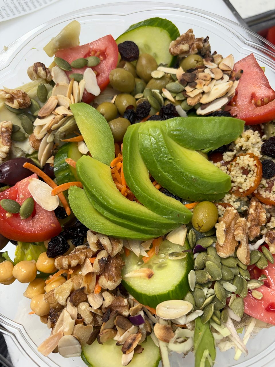 A vibrant salad in a clear container, featuring avocado, tomatoes, olives, nuts, and seeds.