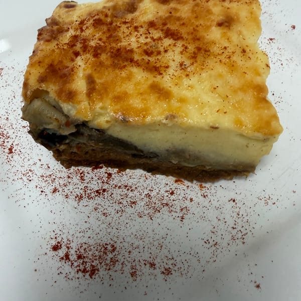 A slice of moussaka with a golden-brown top, sprinkled with paprika, on a white plate.