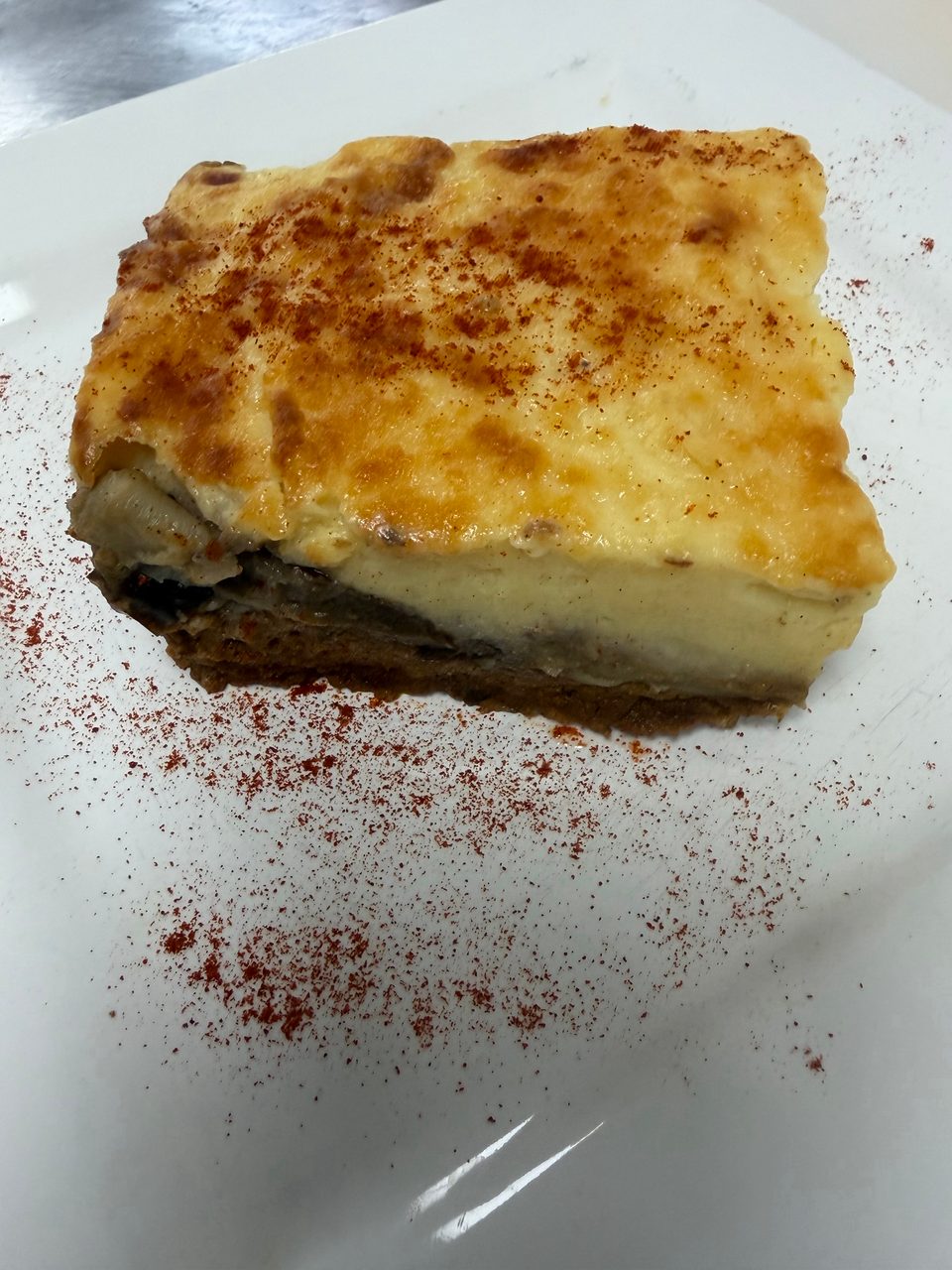 A slice of moussaka with a golden-brown top, sprinkled with paprika, on a white plate.