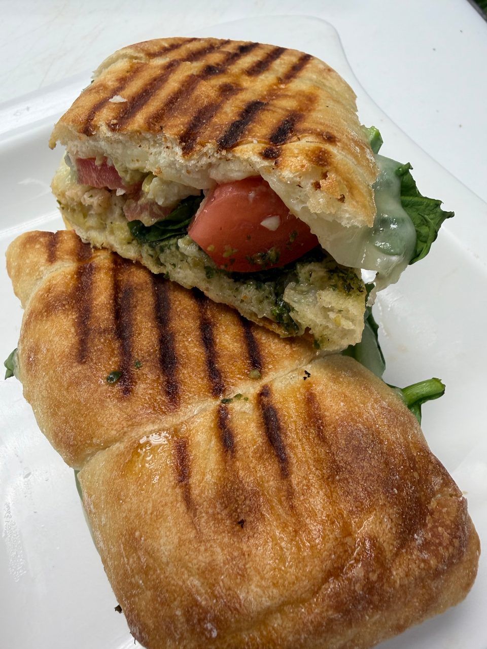 Grilled panini sandwich, cut in half, revealing pesto, tomatoes, and melted cheese on a white plate.
