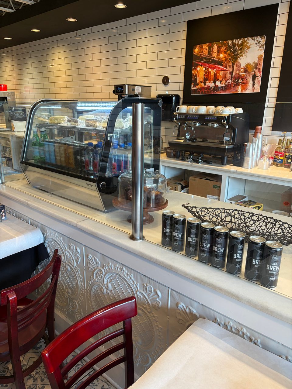 A cafe counter with a pastry display case, coffee machine, and canned cold brew.