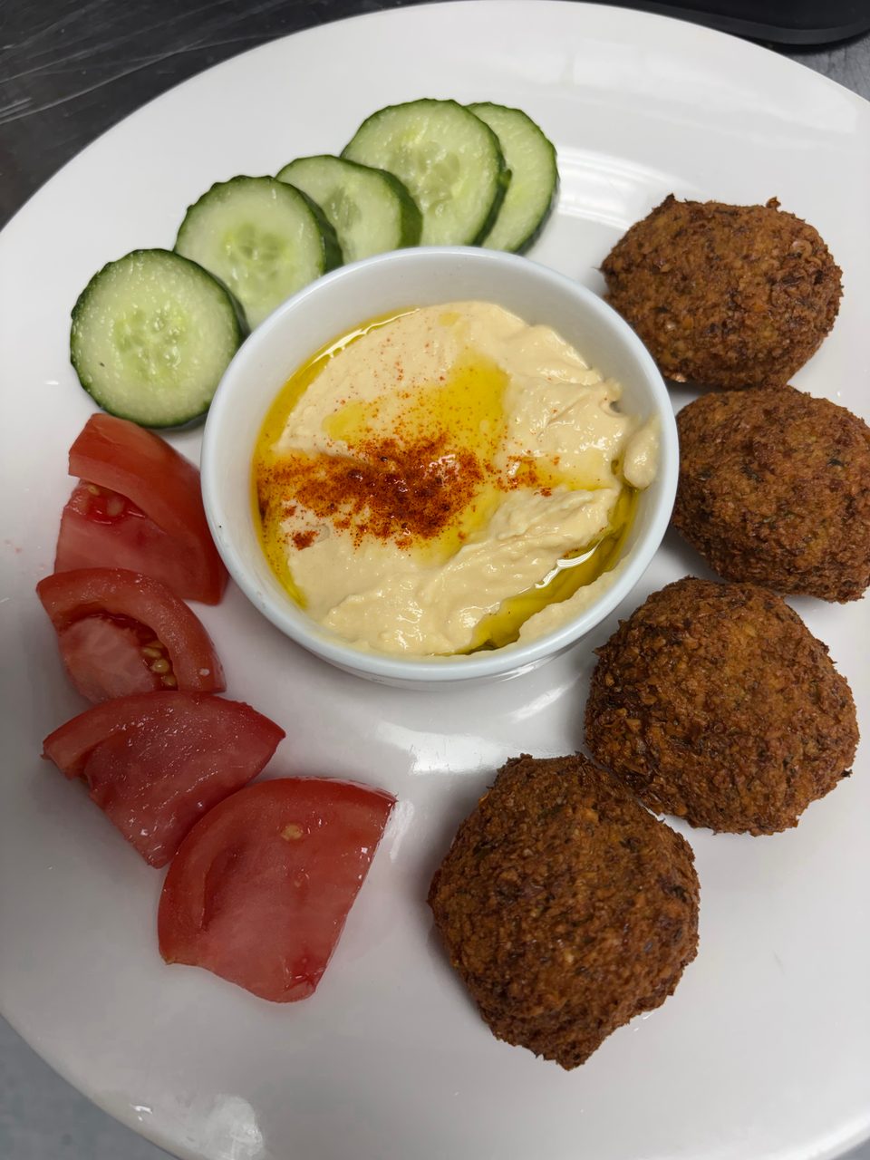 A white plate with falafel, hummus, sliced cucumbers, and tomato wedges.