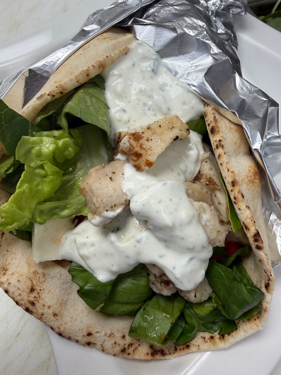 Chicken pita wrap with lettuce, feta cheese, and creamy white sauce, partially wrapped in foil.