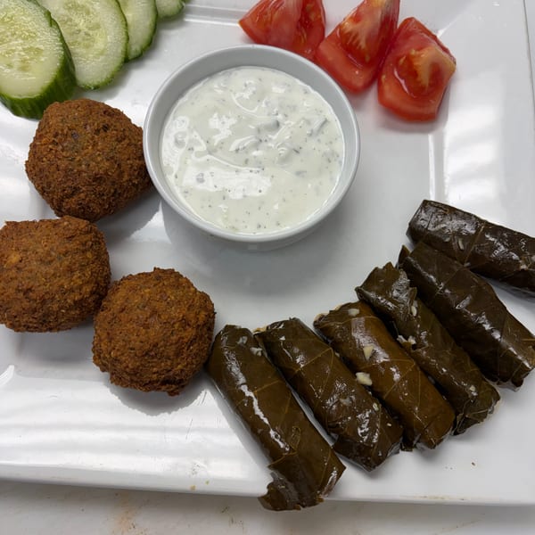 A white plate with falafel, grape leaves, cucumber slices, tomato wedges, and a white dipping sauce.