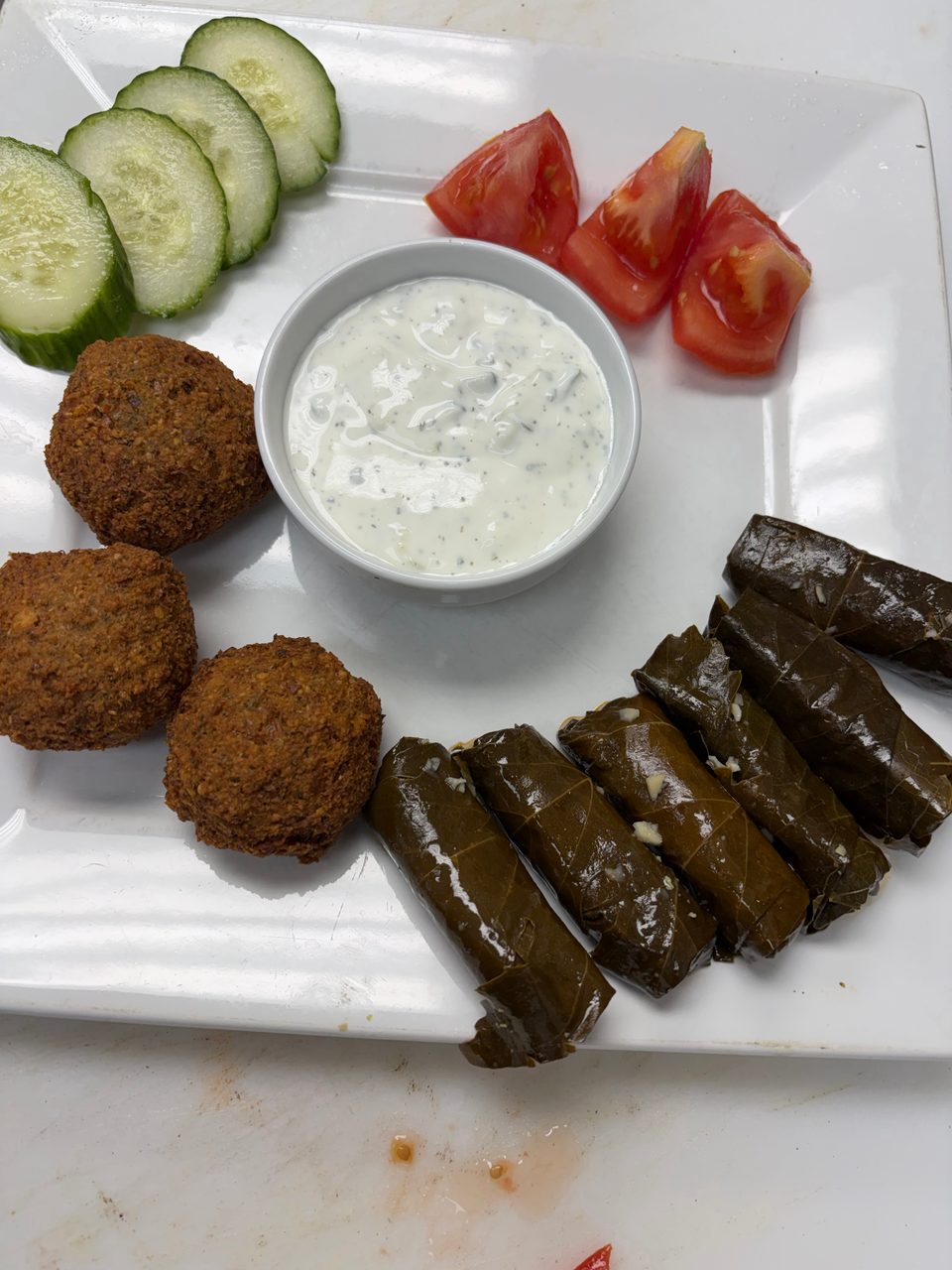 A white plate with falafel, grape leaves, cucumber slices, tomato wedges, and a white dipping sauce.