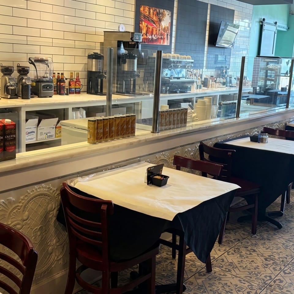 Bright restaurant dining area with black-and-white tablecloths, wooden chairs, tiled walls, and a visible coffee service counter.