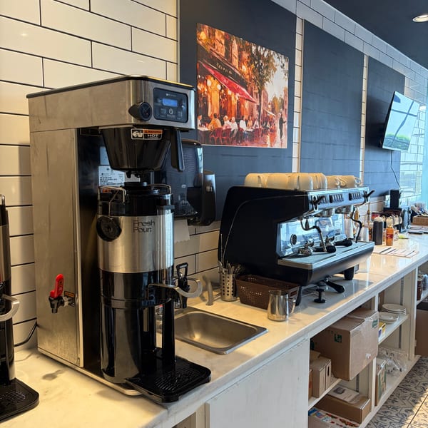 A coffee station with a large coffee maker, espresso machine, and sink against a tiled wall.