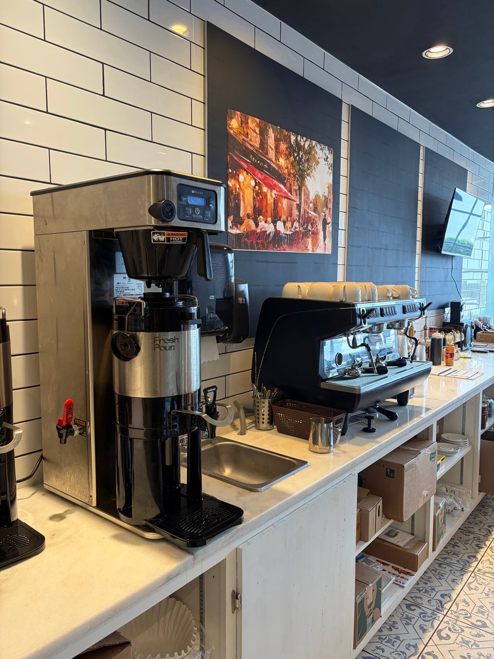 A coffee station with a large coffee maker, espresso machine, and sink against a tiled wall.