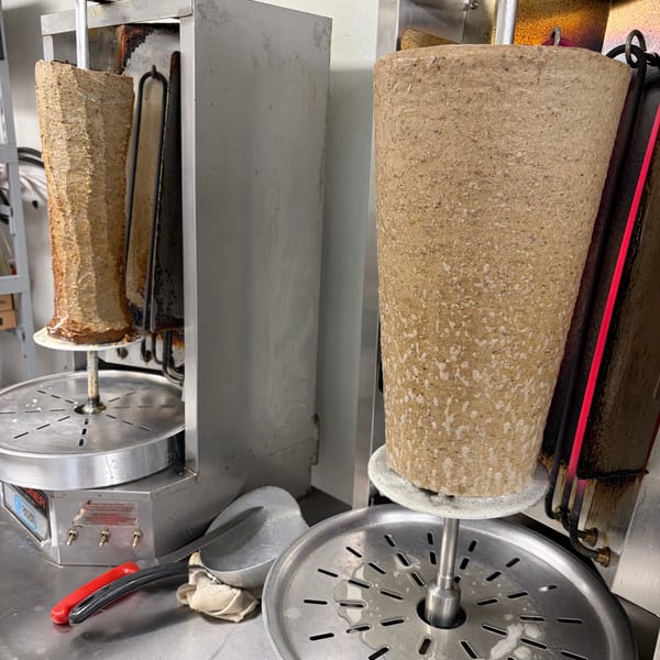 Two large vertical spits of meat for gyros or shawarma cooking on rotisserie machines in a kitchen.