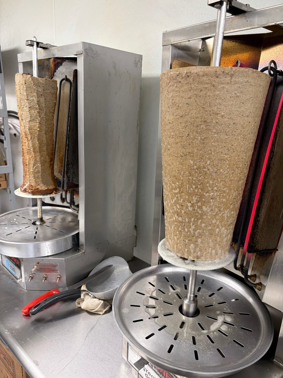 Two large vertical spits of meat for gyros or shawarma cooking on rotisserie machines in a kitchen.