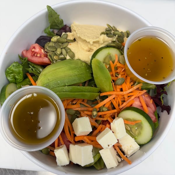 A vibrant salad in a white bowl with hummus, avocado, cheese, vegetables, and two cups of dressing.