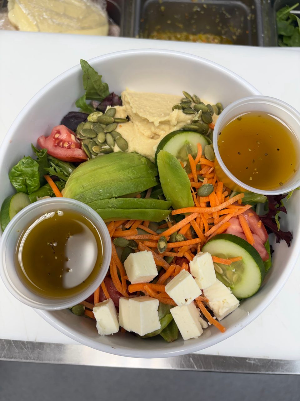 A vibrant salad in a white bowl with hummus, avocado, cheese, vegetables, and two cups of dressing.